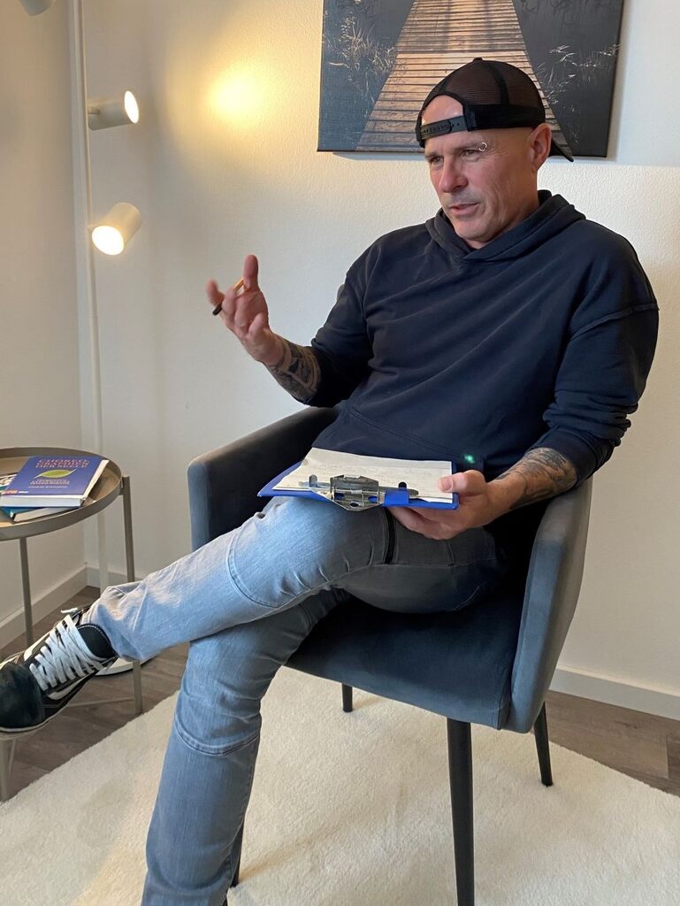 Sepp Kraus sitzt im Sessel und spricht, ein Klemmbrett auf dem Knie